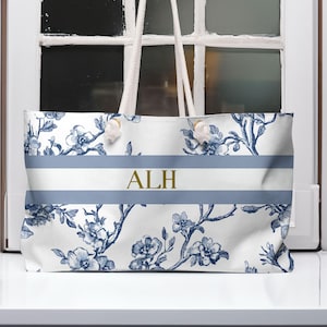 Peut inclure: Sac fourre-tout blanc avec un motif floral bleu et une bande bleu clair au milieu. Les initiales "ALH" sont imprimées en or sur la bande. Le sac a des poignées en corde blanche.