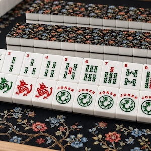 May include: A set of white mahjong tiles featuring green and red ink designs, including numbers one through nine, dragon symbols, joker tiles, and East and South wind characters. The tiles are arranged in rows on a black floral patterned mat.