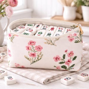 Puede incluir: Un juego de Mahjong crema con un diseño floral. La funda con cremallera está adornada con flores rosas y hojas verdes. El juego incluye fichas blancas con símbolos coloridos, sobre una tela a cuadros.