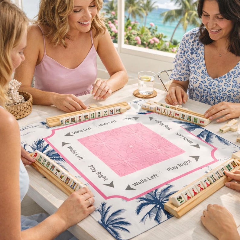 Group of women playing Mahjong on a pink coastal Mahjong mat, styled for game night in a bright beach house setting.