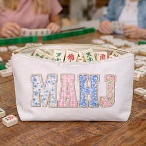 May include: A white canvas zippered pouch filled with mahjong tiles, with the letters "MAHJ" embroidered on the front in floral and gingham patterns. The pouch is on a wooden table with mahjong game pieces and green racks.