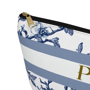 May include: A close-up of a cosmetic bag with a floral and striped pattern. The bag features a white background with blue stripes and floral designs. A gold zipper and the letter "P" are visible.