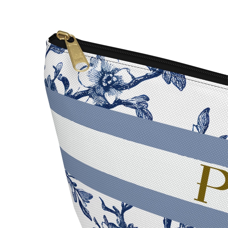 May include: A close-up of a cosmetic bag with a floral and striped pattern. The bag features a white background with blue stripes and floral designs. A gold zipper and the letter "P" are visible.