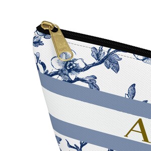 Close-up of a white cosmetic bag with blue floral and striped patterns. The bag features a gold zipper and the letter "A" in gold. The zipper is black and the bag is made of a textured material.