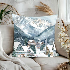 May include: A decorative square pillow featuring a watercolor painting of a snow-covered village nestled at the base of a mountain range. The artwork depicts houses, trees, and a church, all dusted with snow, in shades of blue, gray, and white.