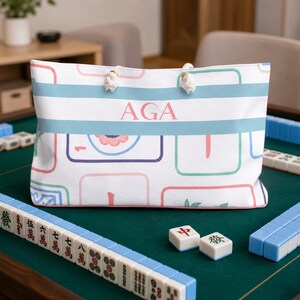 Personalized Mahjong accessory tote bag featuring pastel Mahjong tile illustrations and custom initials "AGA," displayed on a green Mahjong table surrounded by Mahjong tiles and racks in a bright game room setting.
