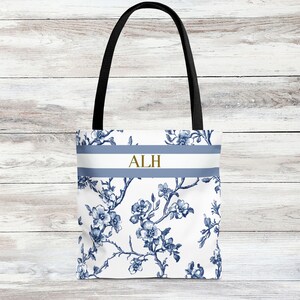 May include: White tote bag with a blue floral pattern and black handles. The bag features a blue and white stripe with the gold letters "ALH". The floral design includes blue flowers and branches.
