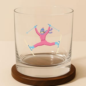 May include: A clear glass tumbler with a whimsical illustration of a skier in a pink jumpsuit, blue hat, and scarf. The skier is mid-air, skis spread, and poles raised. The glass sits on a round, dark wooden coaster.