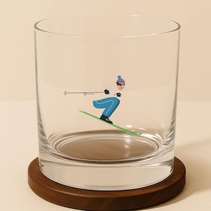May include: A clear glass tumbler with a skier graphic in blue, white, and green. The tumbler sits on a round, dark brown wooden coaster. The skier is wearing a blue hat and goggles.