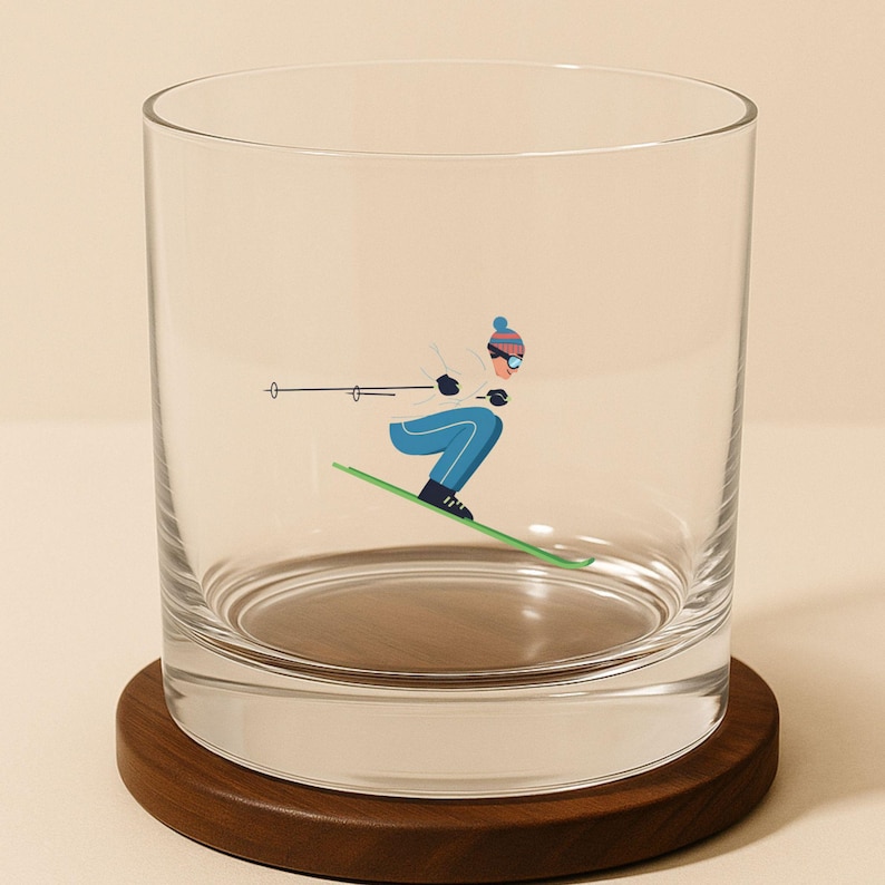 May include: A clear glass tumbler with a skier graphic in blue, white, and green. The tumbler sits on a round, dark brown wooden coaster. The skier is wearing a blue hat and goggles.