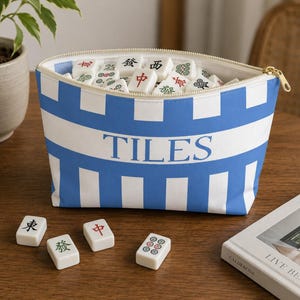 Blue and white striped Mahjong tile bag filled with mahjong tiles sitting on a wooden table with scattered tiles and cozy coastal decor accents.