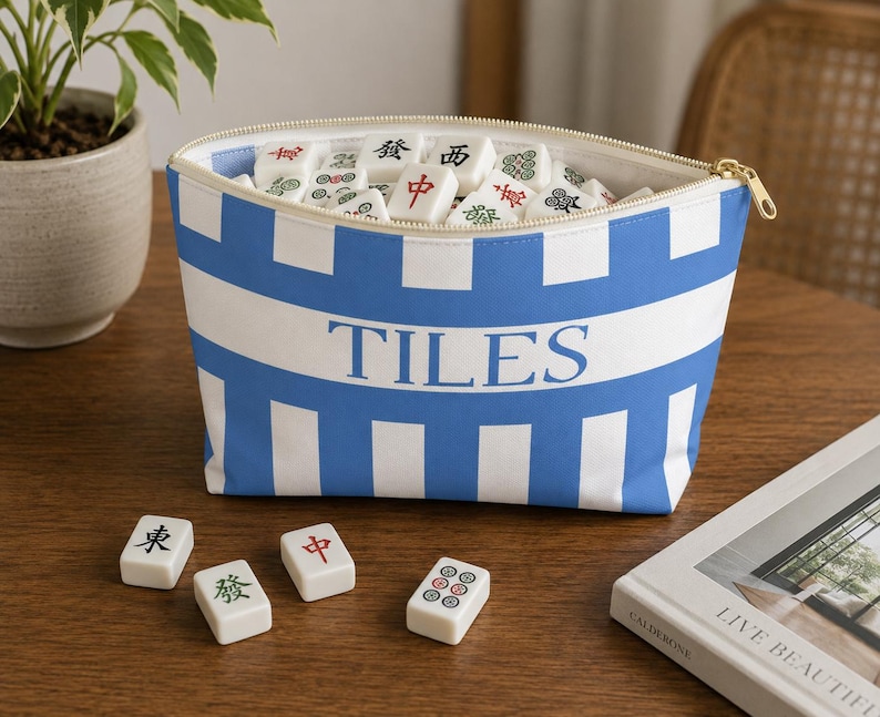 Blue and white striped Mahjong tile bag filled with mahjong tiles sitting on a wooden table with scattered tiles and cozy coastal decor accents.