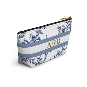 A white cosmetic bag with a blue floral pattern and a gold zipper. The bag features a white stripe with the gold letters "ARD" in the center. The floral design is in a dark blue color.