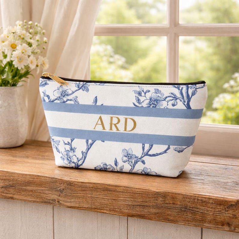 Personalized blue toile accessory bag with gold initials resting on a rustic wooden ledge in front of a country-style window.