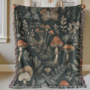 May include: A dark green throw blanket with a botanical design featuring mushrooms, leaves, and foliage in shades of brown, orange, and beige. The blanket has a fringed edge and is held up by a person.