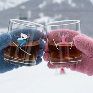 Two ski-themed whiskey rocks glasses clinking outdoors in the snow, held with cozy winter gloves for an après ski moment.