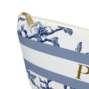 May include: Close-up of a white and blue striped cosmetic bag with a gold zipper. The bag features a floral pattern in navy blue and the letter "P" in gold. The design is elegant and suitable for storing makeup or toiletries.