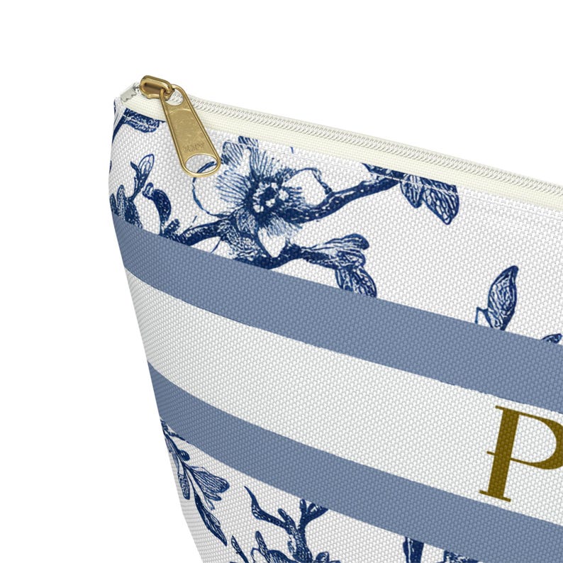 May include: Close-up of a white and blue striped cosmetic bag with a gold zipper. The bag features a floral pattern in navy blue and the letter "P" in gold. The design is elegant and suitable for storing makeup or toiletries.