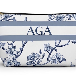 May include: White cosmetic bag with a blue floral pattern and two horizontal blue stripes. The letters "AGA" are centered on the bag. The bag has a gold zipper and a black zipper pull.