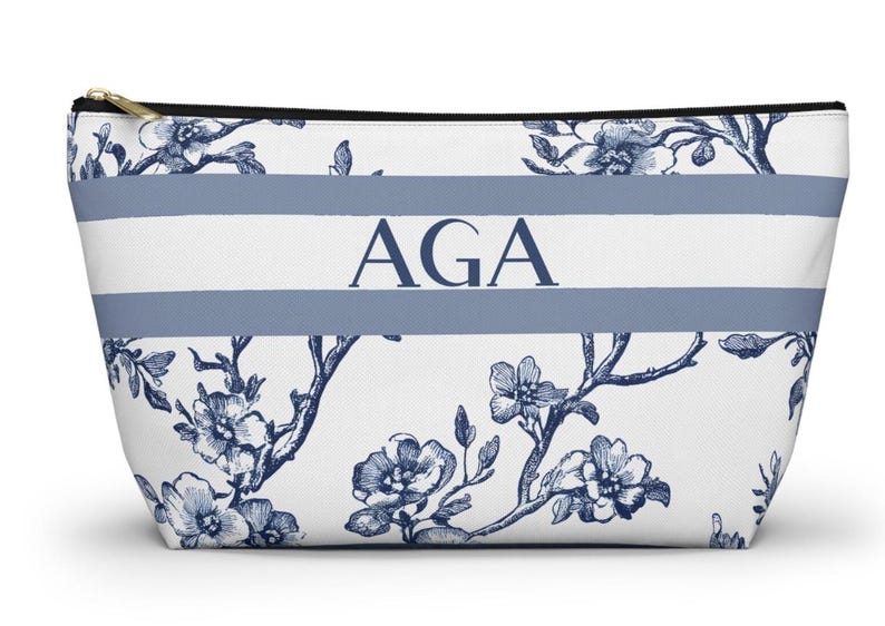May include: White cosmetic bag with a blue floral pattern and two horizontal blue stripes. The letters "AGA" are centered on the bag. The bag has a gold zipper and a black zipper pull.