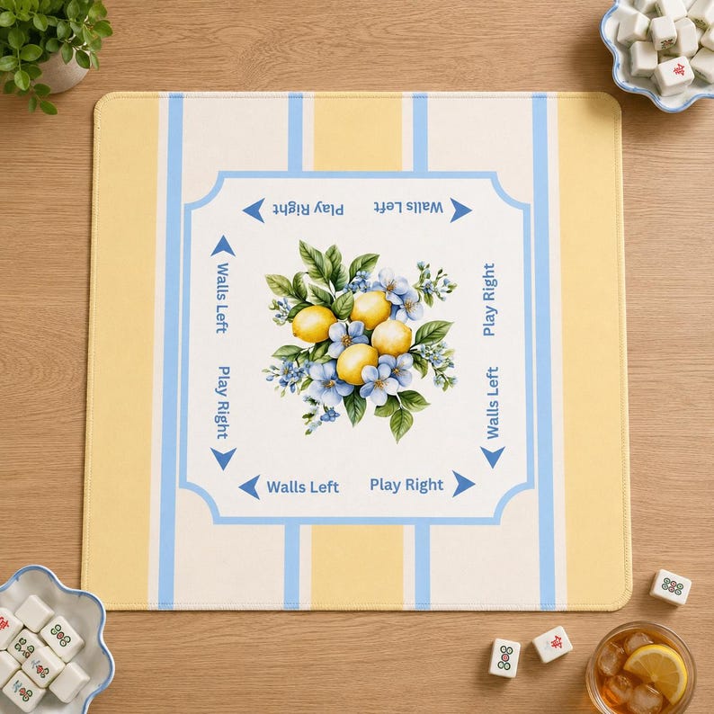 Top-down view of a lemon yellow and blue striped mahjong mat on a light wood table with mahjong tiles, greenery, and iced tea. The mat features a lemon and blue floral centerpiece with directional gameplay markings in soft coastal colors.