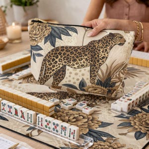 May include: A beige zippered pouch with a leopard print and floral design is held above a Mahjong set. The Mahjong set includes beige tiles with red, green, and blue symbols, arranged on a matching floral mat.