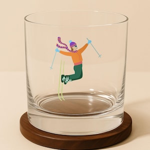 Clear whiskey rocks glass featuring a whimsical skier jumping mid-air with poles raised, shown on a wooden coaster.