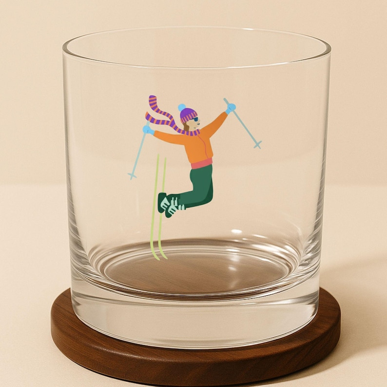 Clear whiskey rocks glass featuring a whimsical skier jumping mid-air with poles raised, shown on a wooden coaster.