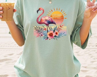 May include: A pale green t-shirt with a flamingo illustration. The flamingo is depicted in pink and purple hues, set against a sunset backdrop with floral and foliage elements. The shirt has short sleeves and a loose fit.
