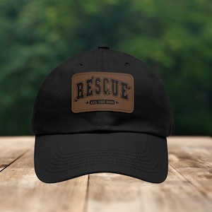 May include: Black baseball cap with a brown leather patch that says "RESCUE ALL THE DOGS" in a vintage style font.