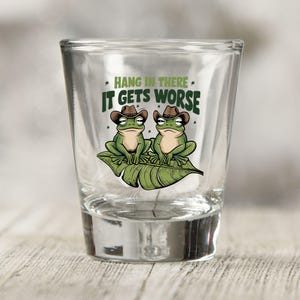 May include: A clear glass shot glass with a green frog design. The frogs are wearing cowboy hats and are sitting on a green leaf. The text "Hang in there, it gets worse" is printed on the glass.