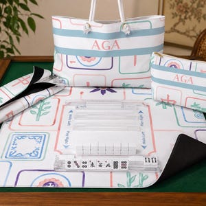 Personalized pastel Mahjong collection displayed on a green felt Mahjong table. The coordinated set includes a 33" x 33" Mahjong mat with colorful Mahjong tile illustrations, a matching monogrammed weekender tote bag with rope handles, a personalized accessory pouch, a rack storage bag holding clear acrylic Mahjong racks, and acrylic racks with Mahjong tiles arranged on the mat.