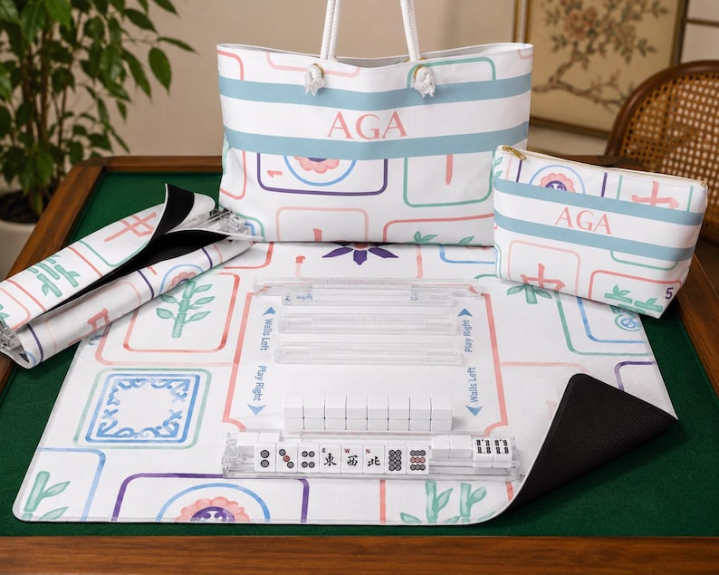 Personalized pastel Mahjong collection displayed on a green felt Mahjong table. The coordinated set includes a 33" x 33" Mahjong mat with colorful Mahjong tile illustrations, a matching monogrammed weekender tote bag with rope handles, a personalized accessory pouch, a rack storage bag holding clear acrylic Mahjong racks, and acrylic racks with Mahjong tiles arranged on the mat.
