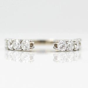 May include: An elegant open-shank ring with a row of sparkling, round-cut diamonds. The diamonds are set in a white metal band, creating a minimalist design. The ring's open design allows it to be worn with other rings.