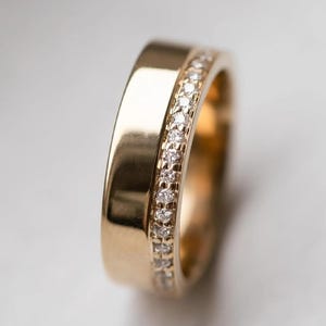 May include: A gold ring with a band of small, clear gemstones set along one edge. The ring has a polished, smooth surface and a classic design. The gemstones sparkle against the gold, creating a luxurious look.