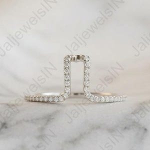 May include: A unique silver ring with a rectangular top, encrusted with small, sparkling diamonds. The ring's design features a modern, open-frame style, with a band of diamonds extending around the finger. The ring is set against a soft, marble-like background.