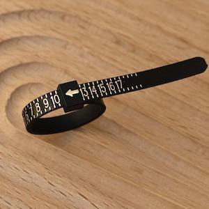 May include: A black ring sizer tool with white numbers and markings. The tool is made of a flexible material and is used to measure ring sizes from 7 to 17. The tool is lying on a wooden surface.