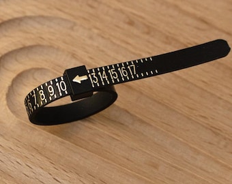 May include: A black ring sizer tool with white numbers and markings. The tool is made of a flexible material and is used to measure ring sizes from 7 to 17. The tool is lying on a wooden surface.