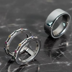 May include: Two silver-toned rings featuring small, multi-colored circular gemstones set along the outer band, positioned next to a single, wider, plain silver-toned band with a smooth, polished finish and a reflective surface.