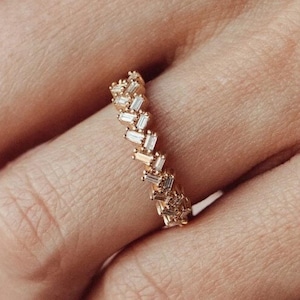 May include: A gold ring featuring a chevron pattern, adorned with small, rectangular gemstones. The ring is on a finger, set against a soft pink backdrop.