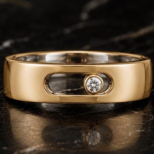 May include: A gold band ring featuring a horizontal, elongated oval cutout on the front surface. Inside this cutout, a single, small, round, brilliant-cut white diamond is set in a circular bezel, positioned toward the right side of the opening.