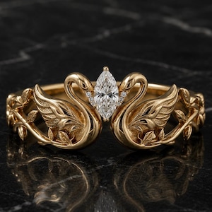 May include: A gold ring featuring two sculpted swans facing each other with wings spread. A marquise-cut clear gemstone is set between the swan heads. The band is detailed with embossed leaf patterns extending from the swan wings along the sides.