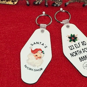 May include: Two white Christmas-themed keyrings with silver key rings. One keyring features Santa Claus and the text "SANTA'S WORKSHOP." The other keyring has the address "123 ELF RD NORTH POLE 88888."