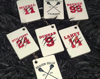 May include: Six white rectangular keychains with red and black lettering and graphics. The keychains feature the names McCall, Raeken, Stillinski, Dunbar, Lahey, and Beacon Hills Lacrosse. Each keychain has a claw mark design.