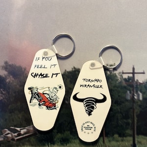May include: Two white keychains with black text and images. The first keychain says "If you feel it chase it" and has a cartoon image of a person driving a red car. The second keychain says "Tornado Wrangler" and has a black silhouette of a bull with a tornado inside. Both keychains have a silver ring.