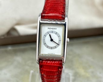 May include: A rectangular silver wristwatch with a white face and black hands. The watch features the "Yves Saint Laurent" logo and a vibrant red leather band. The watch is displayed on a clear stand.