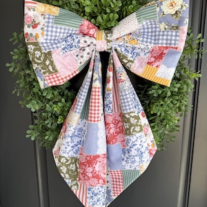 May include: A large decorative bow made of patchwork fabric featuring a variety of floral, gingham, and striped patterns in shades of blue, pink, yellow, green, and white, centered over a circular wreath of small green leaves.