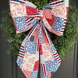 May include: A large, patriotic bow with a patchwork design in red, white, and blue. The bow features various patterns, including floral prints, stripes, and stars, and is attached to a green wreath, perfect for door decor.