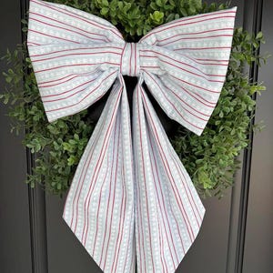 May include: A decorative wreath featuring a large bow made of fabric with a pattern of red stripes and white stars on a light blue background. The bow is centered on a green, leafy wreath, suitable for seasonal home decor.