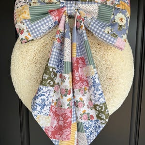 May include: A large decorative bow made of patchwork fabric featuring a variety of floral, gingham, and striped patterns in shades of blue, green, pink, yellow, and white. The bow has two long, pointed tails hanging down over a round, textured cream base.